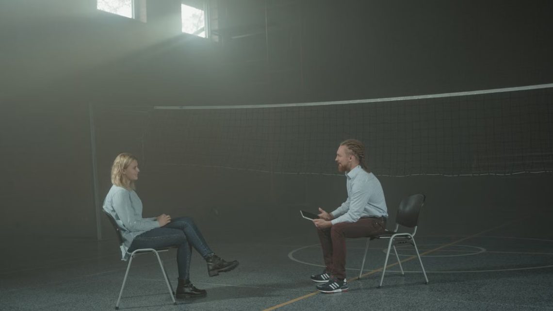 A man and a woman engage in a therapy session in a smoky indoor setting, sitting apart in a large room.