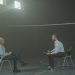 A man and a woman engage in a therapy session in a smoky indoor setting, sitting apart in a large room.